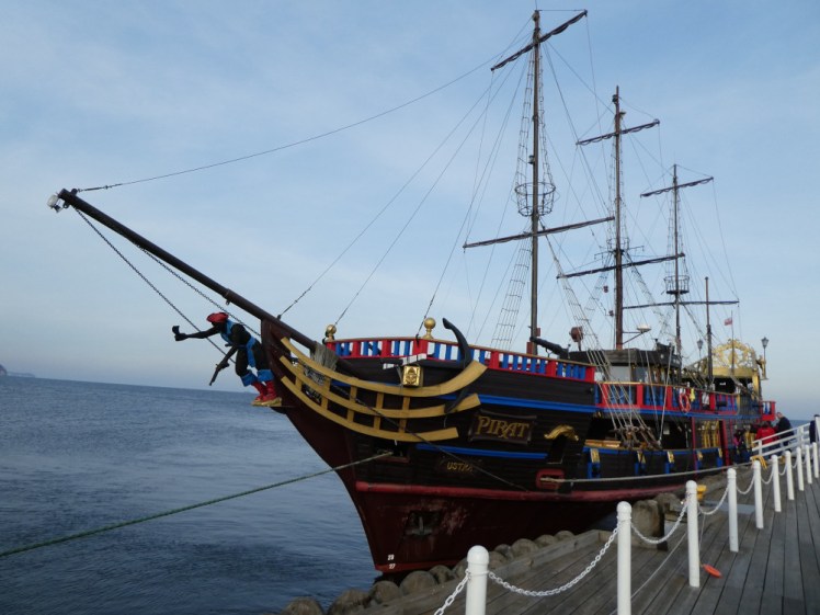 A colourful pirate ship tied up alongside the port. There are no sails on her three masts and the railings are red and blue.