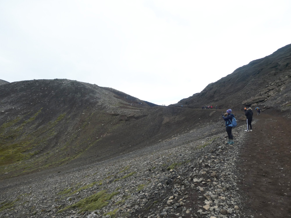 A rocky gravelly slope going up the side of a mountain while tourists stop to take photos across the plain to the left.