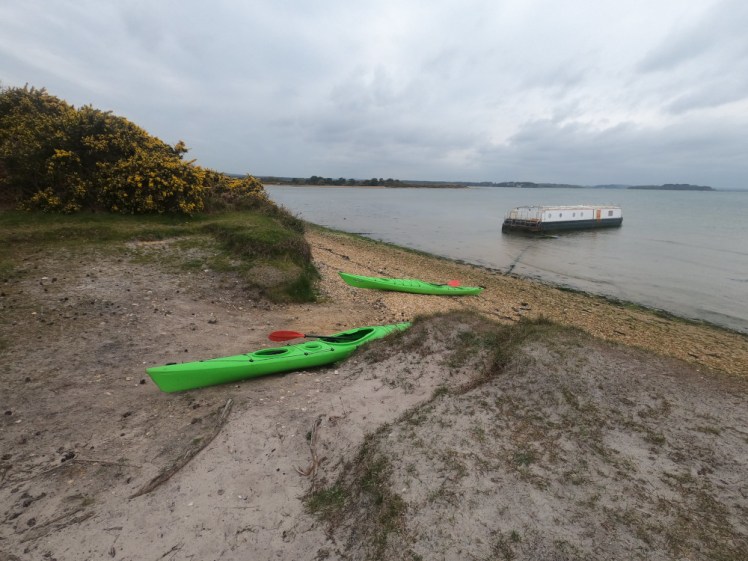 Two long green kayaks on the beach. Well, there's a strip of orange pebbly beach and then a length of greyish sand which is actually scrubby dune. The sky and sea are both pretty grey.