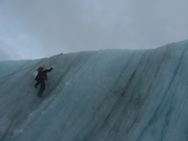 Me, wearing crampons and wielding ice axes climbing a wall of blue streaked ice.