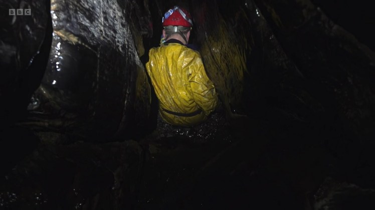 Caving in Cwm Dwr. A caver in a yellow PVC suit and red halmet slithers between shiny rocks, his back to the camera. I think there's a stream under him too.