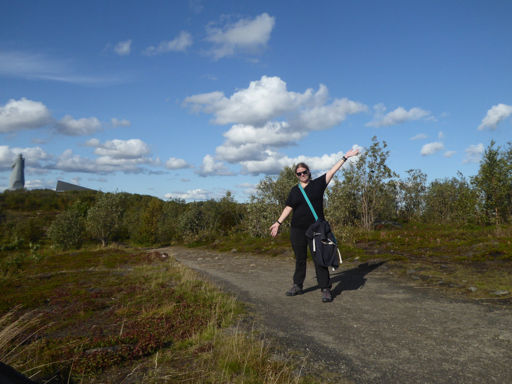 A timer selfie of me in the middle of an Arctic wilderness in summer. The trees are scrubby, the grass is more moss than anything else, the sky is blue and there's a 116ft concrete soldier statue behind me. I'm wearing a black t-shirt and a teal cross-body bag which is holding up a navy hoodie. I'm standing with my left arm up and my right arm down, like it's the first chance I've had to take a proper selfie in nearly two weeks.