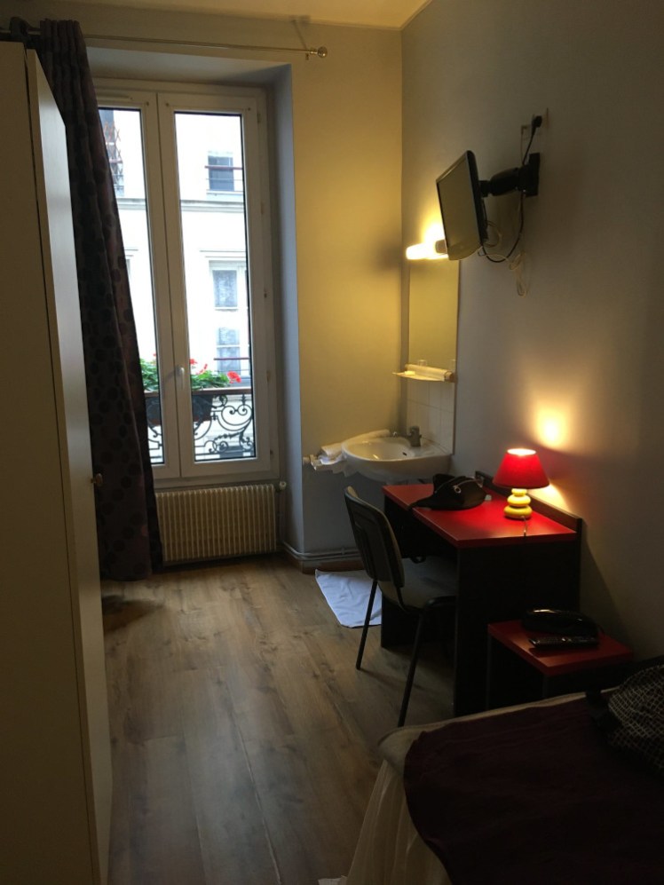 A dingy hotel room near Gare du Nord in Paris. It's a narrow room, lit only by a small lamp and a light over the sink. The door doesn't fit properly and it smells weird.