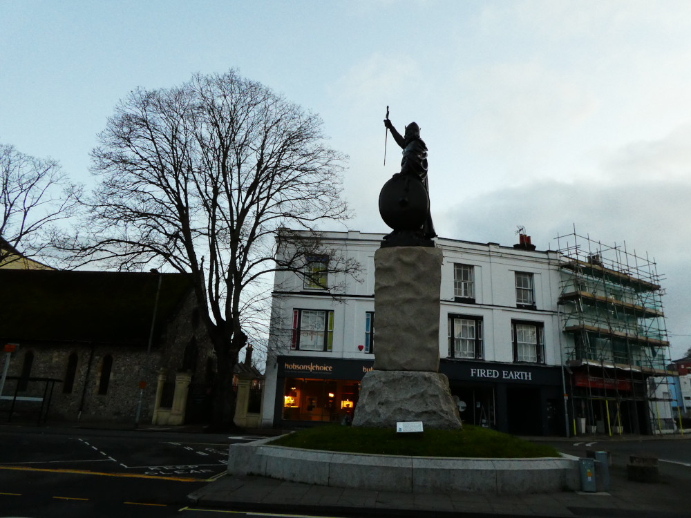 The statue of King Alfred the Great in Winchester, holding a sword and shield on top of a rough stone pillar, silhouetted against the early morning bright blue-white sky.
