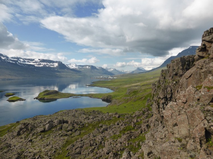 The view across a fjord reflecting the blue sky patched with cloud. On the opposite side, the mountains are steep with patches of snow. On this side, the mountains are basaltic and their slopes covered in grass.