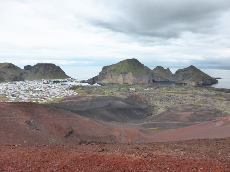 Sitting on the top of Eldfell, looking down into the crater, which is open on the opposite side because its edge broke off in 1973, and a lava field sweeping down to the sea. On the other side of the harbour are craggy orangey cliffs. The volcano and its crater are bright red rock with blacker rock lower down.