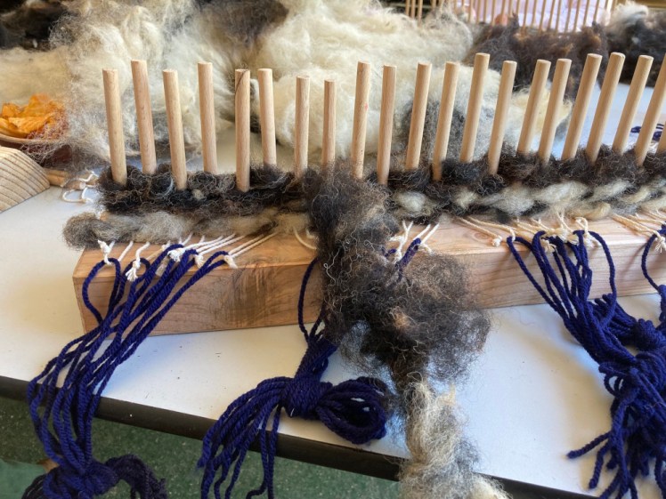 Starting to weave - maybe four layers of raw fleece woven around the pegs of the loom. There are now lengths of navy yarn tied onto each of the string loops and these are tied together in bundles to keep them out of the way.
