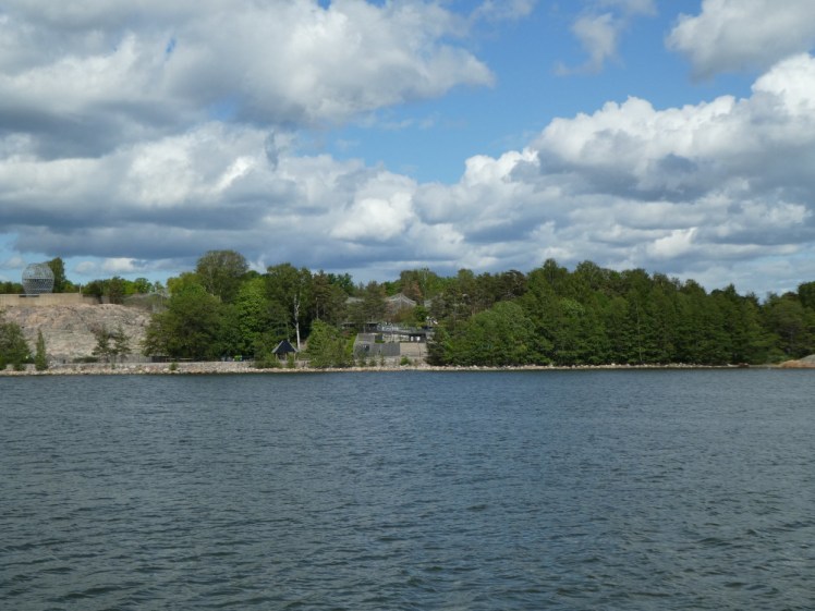 Korkeasaari, the zoo island, seen from the water. A lot of the island is covered in trees and you can see patches of bare rock and sides of enclosures.