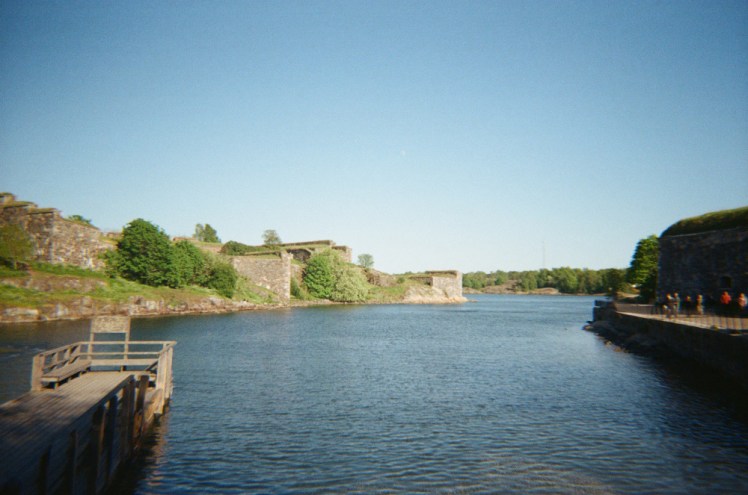 An inlet between islands on Suomenlinna, lots of deep blue water and on each side are thick walls and trees. The sky is very blue.
