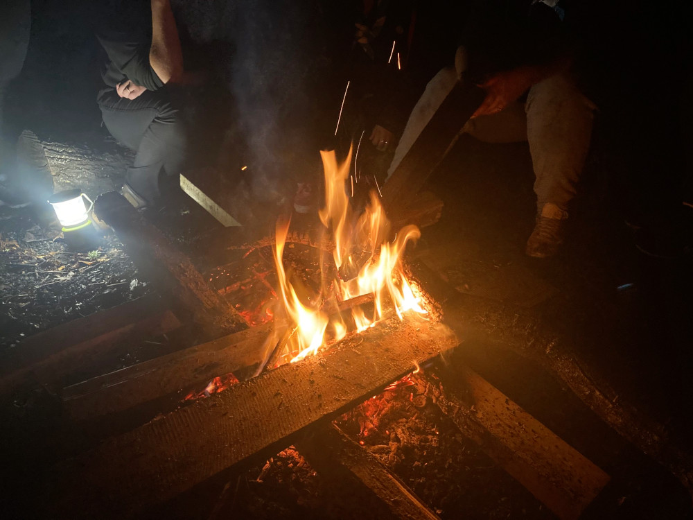 A fire surrounded by damp planks. People are sitting around the edge of the campfire circle - they look very close and they are but they're not dangerously close.