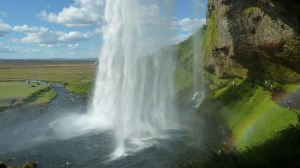 A large lacy waterfall lands in a blue plunge pool, seen from the cave carved out behind the waterfall. This is Seljalandsfoss. Behind the waterfall, a rainbow arcs through the spray. On the other side, a stream runs away from the waterfall and grassy plains stretch into the distance.