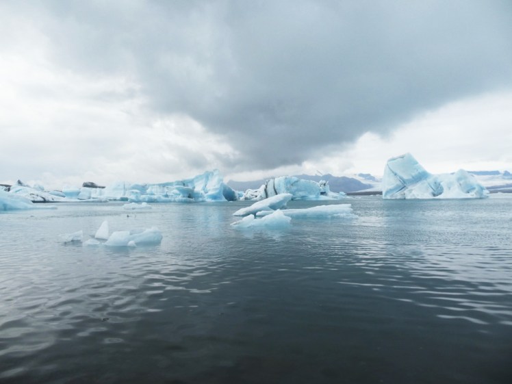 Jökulsárlón, the glacier lagoon, with a dramatic cloud over it, and striped blue, white and black icebergs floating in the dark blue water.