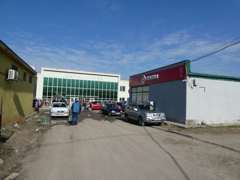 A junction of Tserovani streets with the school, which has a large glass front, facing a small assortments of shops, including local grocery chain Magniti.
