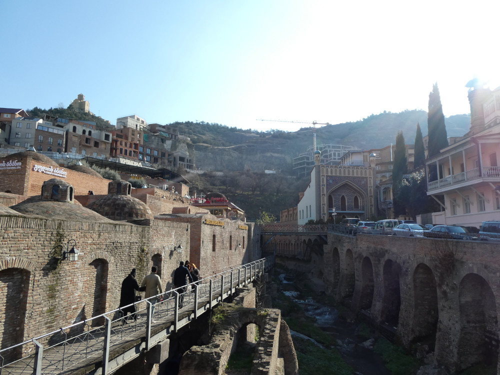 A narrow shallow gorge with a hot river down the middle. The sides are now brick and above and behind each brick wall is a sulphur bathhouse. You can see the blue-tiled frontage of Chreli Abano on the right and the brick front of King Erekles on the left.
