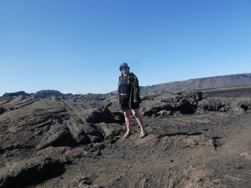 A timer selfie of me standing on a rock in the middle of a lava field. The sky is very blue and I'm wearing sandals, shorts & a crop top, although I have an open shirt over it because it's a little bit breezy.