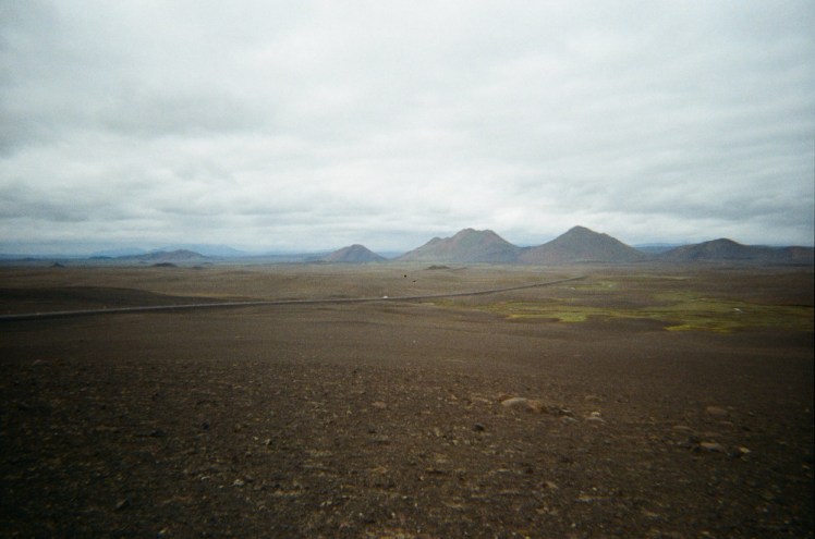 A gravelly lava field with three mountains on the horizon, under a cloudy sky.