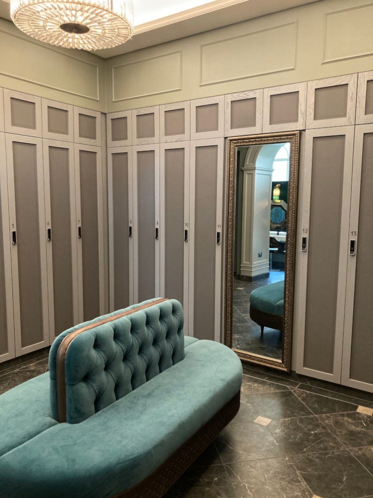 The changing rooms. Light-coloured lockers with darker beige-grey insets reach up to about six feet and there's an off-green sofa-thing in the middle.