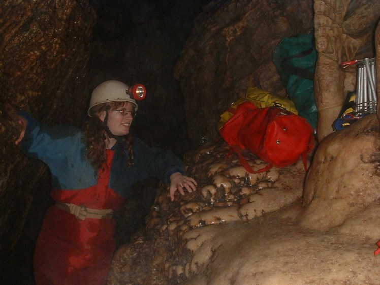 A photo of me from 2004 in a limestone cave. The photo is a bit dark - cave photography was not what it is now. I'm wearing a white helmet with a chunky lamp sticking out and a wet red and blue caving suit. Piled up on the rock opposite me are several tackle bags and a rolled-up electron ladder.