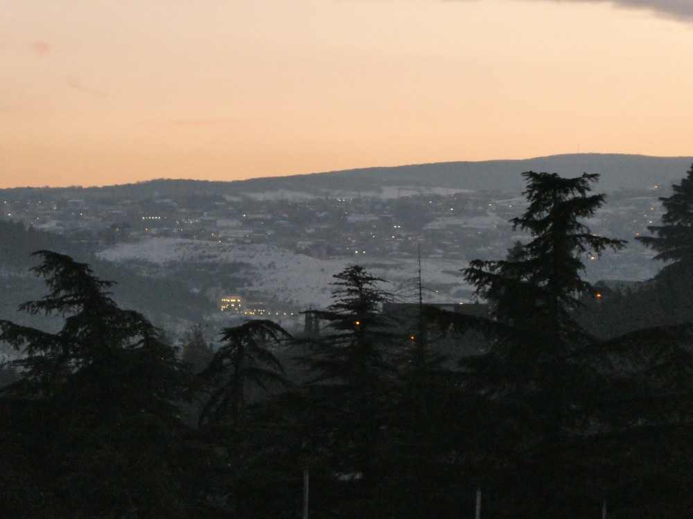 The hazy view across from the entrance to the park towards a mountain covered in equal parts in snow and buildings with faint lights in the window. At this level of zoom, the sky is a peachy orange.