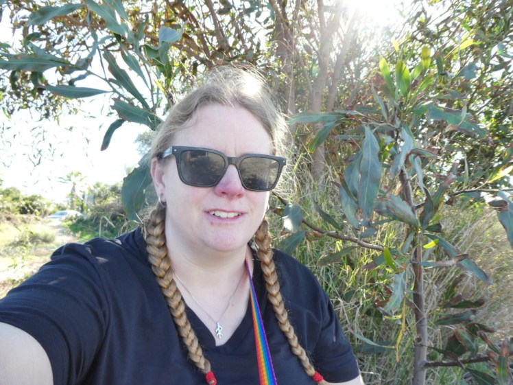 Selfie under a tree by the salt lake. I'm wearing sunglasses and a black t-shirt, with my hair in plaits and the sun trying to push its way into the picture.