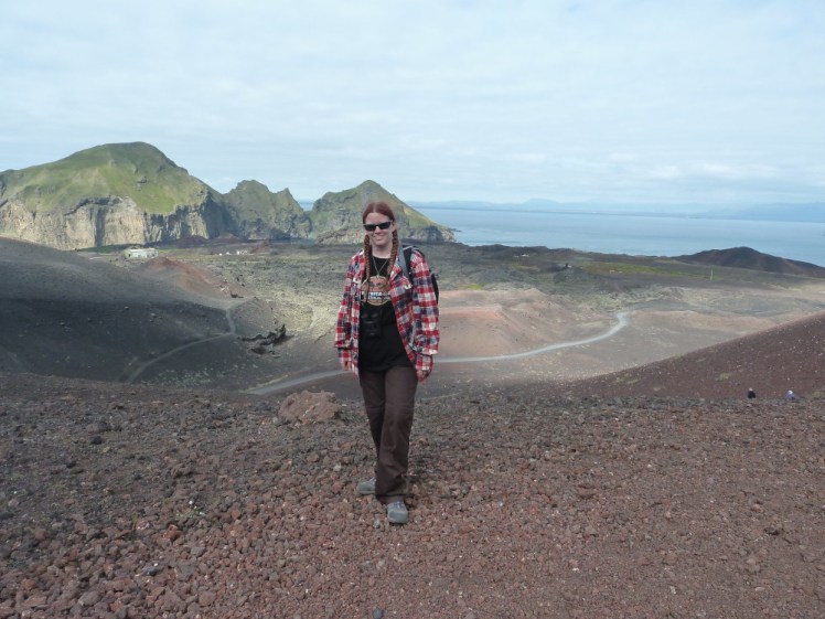 Me, in a red checked shirt and brown trousers, standing in the Eldfell crater, with a reddish volcanic field behind me and cliffs and the sea in the distance.