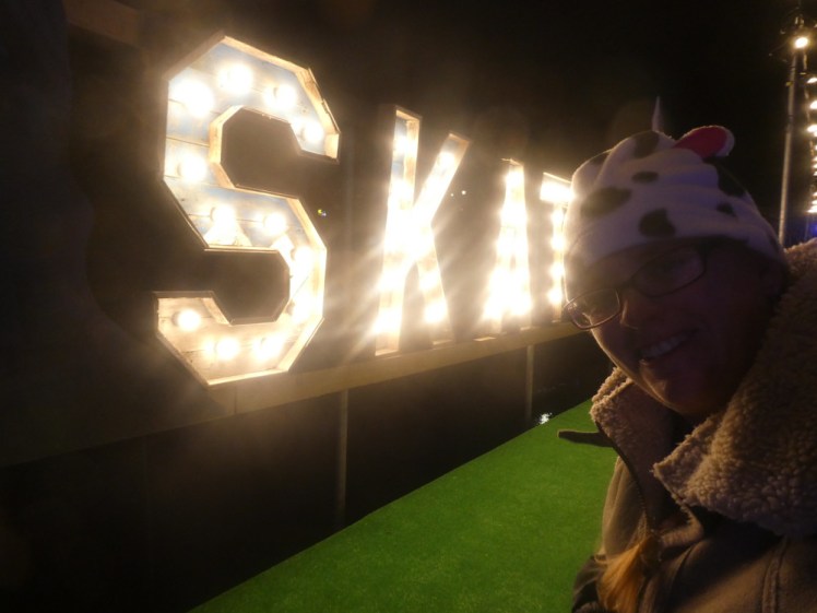 A selfie backlit by huge illuminated letters saying SKATE at the side of the rink. You can't actuallys ee the E because my head is in the way; I'm in silhouette because of the lights in the letters. You can't actually see that I'm standing on the ice either.