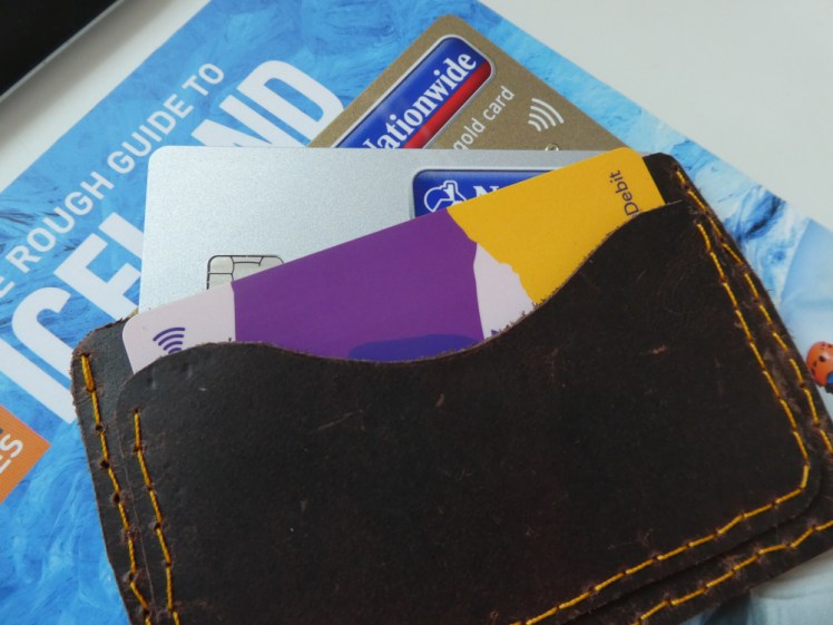 A handmade leather card-holder with wonky orange stitching. Poking out of it are a purple and yellow debit card, a gold (brownish!) credit card and a silver one.