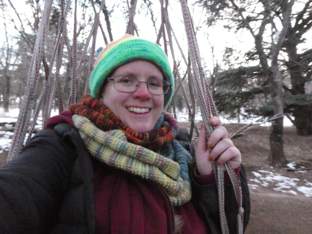 Me in a colourful hat and scarf grinning in a knotted rope hanging hammock-like chair. Behind me, you can see snow on the ground.
