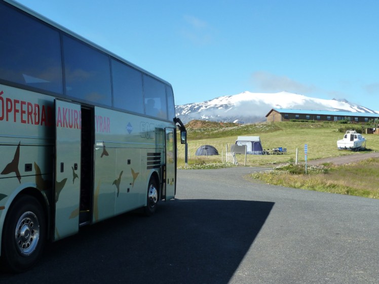 The snow-capped mountain again, this time framed by a sage-green tour bus and a long low wooden building on a small hill.