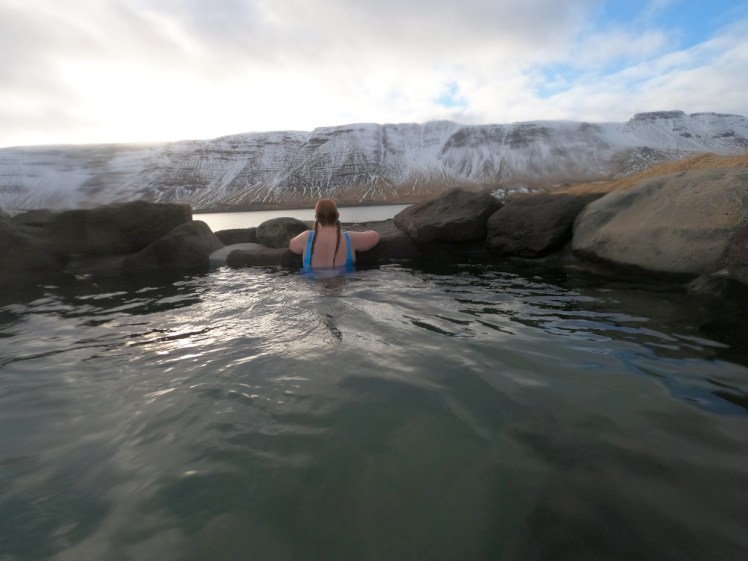 My back, with two red plaits hanging down and the bright blue straps of my polar bear swimsuit. I'm sitting at the edge of the Beach Pool, arms on the side, elbows out, looking out at the view. From here you can see the sun on the water in the pool, the rocks surrounding it and then the ridge of snowy mountains directly opposite, with just a slither of fjord visible.
