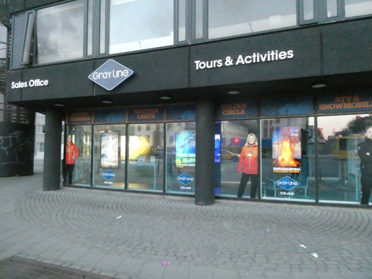 Grayline's downtown office in 2022, a building that seems to be held up with stilts and the office behind the stilts so that the upstairs overhangs the downstairs. Grayline's windows are full of pictures of their staff in orange outdoors jackets, eruptions, Northern Lights and lots of logos.