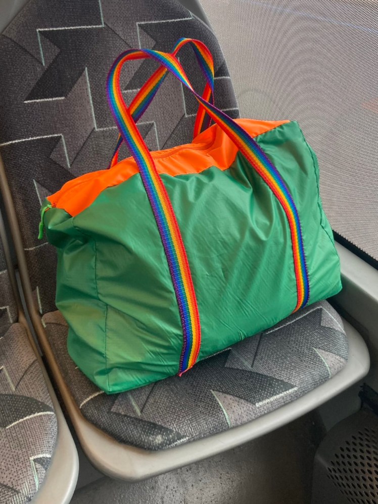 A rectangular green nylon bag with an orange panel in the top and rainbow straps around the whole bag, sitting on a bus seat.