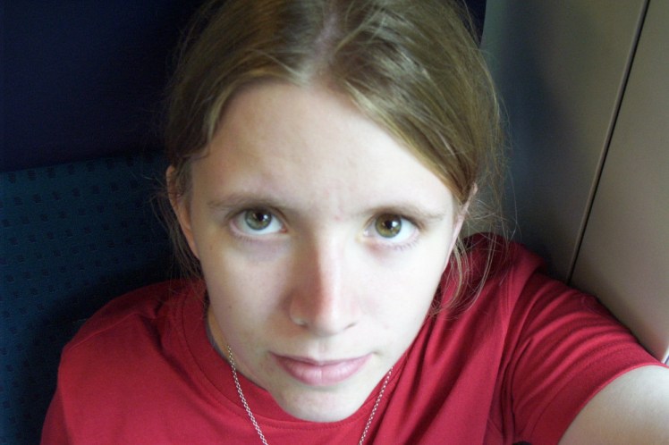A selfie taken, MySpace-style, from above, making my face much narrower at the bottom than the top. I've deliberately taken off my glasses and if you look closely, you'll see that although my eyes are vaguely green, my right one is slightly bluer than the left, which is slightly browner. I'm wearing a red t-shirt and you can make out the green and blue seats of the long white Swiss trains.