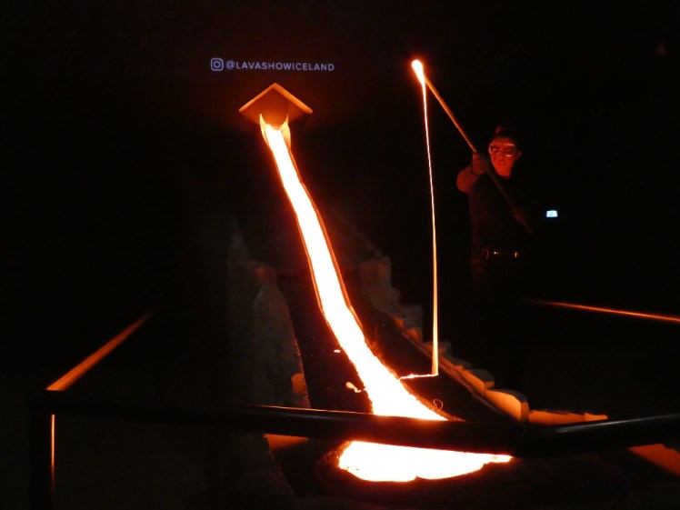 In a darkened theatre, lava flows down a steel slide, illuminating the room in orange although it glows white itself. A man with a steel pole has scooped up some lava which is dangling from the pole like thick custard.