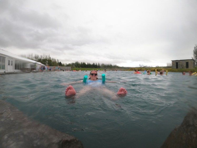The Secret Lagoon: Me, floating on a pool noodle with my feet pointing at the camera. The pool is a large but reasonably shallow pool with grass on three sides and a long white building on the left.