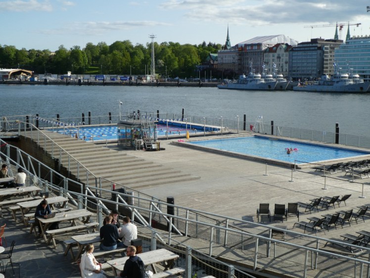 A large wooden deck protruding into a harbour. On the other side of the harbour are some buildings on the right and a lot of thick trees on the left. Set into the deck are two bright blue swimming pools.