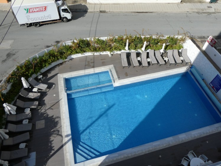 The hotel pool as seen from a balcony four floors up. It's rectangular, bright blue, with the shadow of the hotel across the corner, and separated from the road by a thin fence.