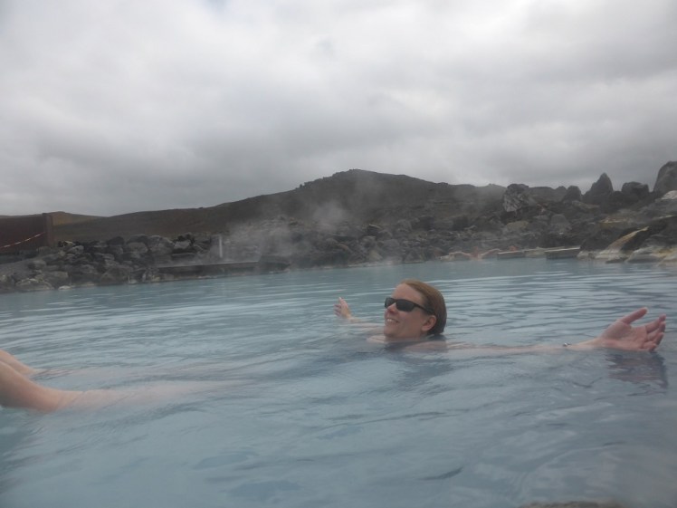 Me, floatingo n my back in a quiet corner of Myvatn Nature Baths with my arms outspread. The water is bright blue and steaming. Behind me, the lagoon is ringed with boulders and behind that is the slope of an active volcano.