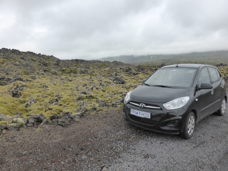 A small black Hyundai i10 parked next a very rough lichen-covered lava field.