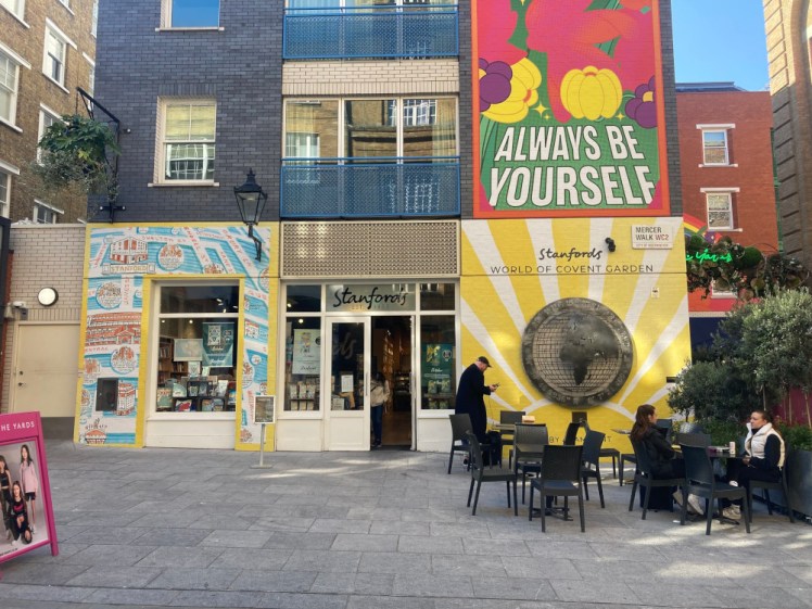 Stanford's map and book shop, a shop front almost invisible behind three big murals. The one on the left is a big yellow and blue map of the area, the one on the left is a big yellow sunburst with a brass plate of the globe in the middle and above that is a bright pink one with fruit declaring "always be yourself".