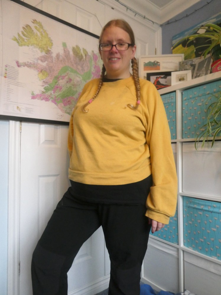 Me in a cropped mustard-yellow jumper standing in my Iceland corner. My hair is in plaits and I'm wearing black hiking trousers with a longer black t-shirt camouflaged but sticking out under the jumper.