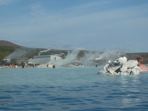 Svartsengi, the geothermal power station, as seen from the Blue Lagoon. They sky is blue, the water is even bluer and in a cloud of steam, a grey building, stubby cooling towers and something silver can be seen.