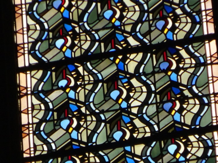 Stained glass high up in the clerestory where you'd only notice the intricate patterns with a good zoom. It's abstract, in shades of yellowy green and blue, with wiggly blue lines separating the patterns into something resembling columns.