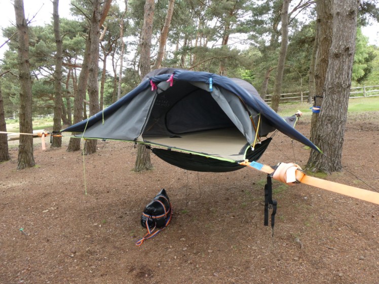 A triangular tent on a trampoline-like platform suspended between three trees, with its black waterproof cover folded back and its inner rolled up to show the empty interior.