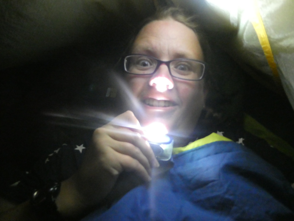 A selfie in a blue sleeping bag in the tent in the dark. I'm holding my headtorch under my face to light from beneath so I look scary like we did in junior school. It doesn't work. I look ludicrous, not scary.