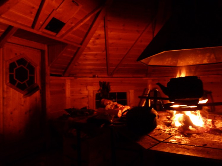Inside the barbecue hut, which is lit only by the roaring fire in the firepit in the middle. There's an iron kettle on a stand silhouetted by the fire.