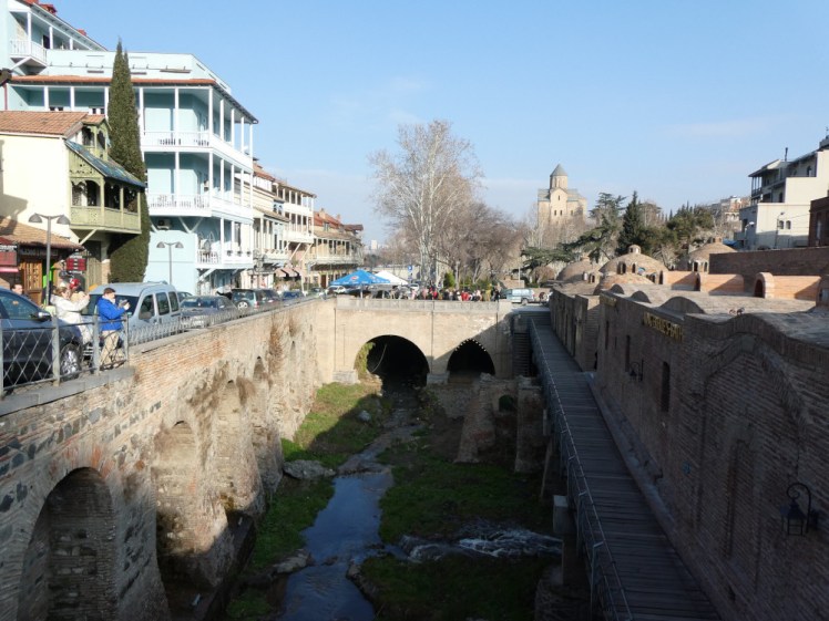 A gorge shadowed in the lowish sun. There are brick walls, the sides of bathhouses, on each side and a brick bridge at the end. In the bottom is a small stream. The fact that the grass around it is neon green tells you how warm it is.