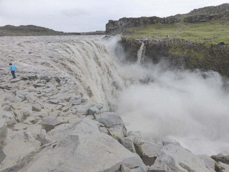 A huge white waterfall falls into a chasm sending up a cloud of spray. The east side, nearest the camera, is just broken grey rocks - you can't tell where the rocks end and the water begins. The west side is visible, a sloping green meadow with a low cliff at the top.