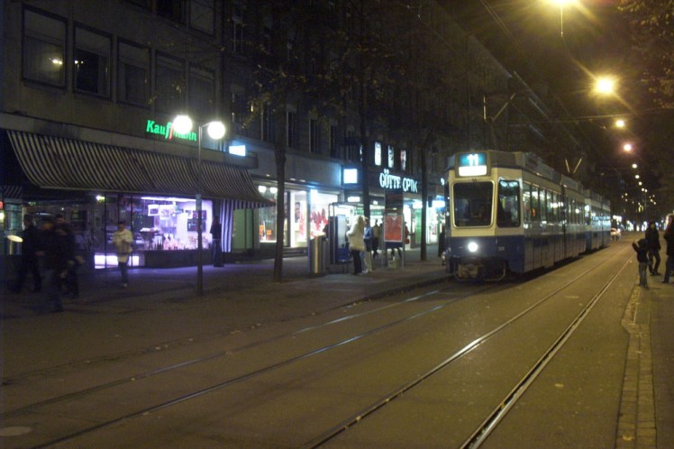 Tram in Zurich by night, running down a street with bright yellow lights overhead.