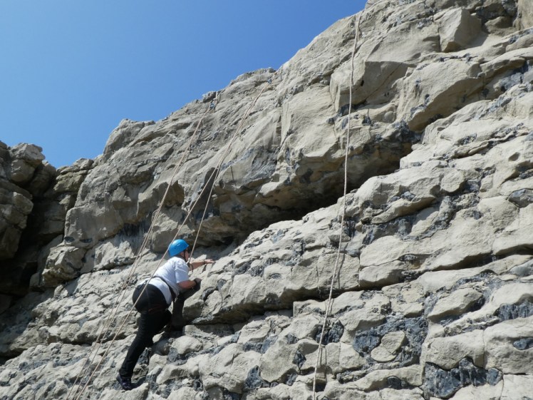 Me, in a blue helmet and climbing harness, climbing up a stratified limestone wall.
